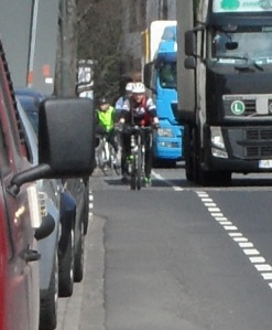 Drei Radler fahren - von vorn fotografiert - hintereinander auf dem schmalen Schutstreifen auf der Leipziger Straße; links von ihnen sind parkende Autos zu sehen, rechts fahren Lkws vorbei.